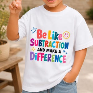 May include: A white short-sleeved t-shirt featuring the colourful text "Be Like SUBTRACTION AND MAKE A DIFFERENCE" in various fonts and colours, accompanied by a blue star, a yellow smiley face, a yellow sparkle, and blue asterisks.