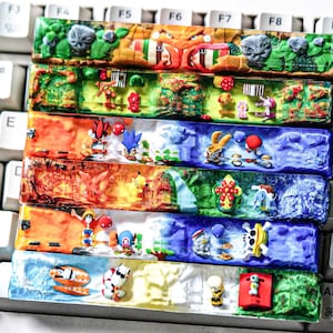 Peut inclure: Une collection de six barres d'espace de clavier en résine rectangulaires, chacune présentant des dioramas tridimensionnels complexes et multicolores de divers personnages animés et paysages, posées horizontalement sur un clavier d'ordinateur blanc.
