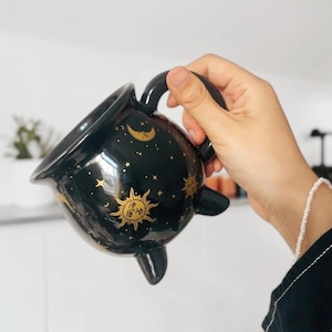 Puede incluir: Una taza de cerámica negra con forma de caldero que presenta un patrón celestial dorado de soles, lunas y estrellas. La taza tiene un cuerpo redondeado, un asa curva y tres pequeñas patas en la base. El acabado brillante refleja la luz.