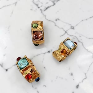 Könnte beinhalten: Drei goldfarbene Ringe mit einer texturierten, unebenen Oberfläche. Jeder Ring ist mit mehreren eingebetteten, mehrfarbigen Cabochon-Steinen in Grün, Bernstein, Blau und Rot besetzt. Die Ringe haben eine klobige, unregelmäßige Form.