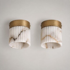 Op de afbeelding: Twee cilindrische plafondlampen met een geribbelde behuizing van wit marmer met natuurlijke grijze en gouden aders. Elke lamp is afgewerkt met een ronde kap van geborsteld messing, wat zorgt voor een strakke en gestructureerde uitstraling.