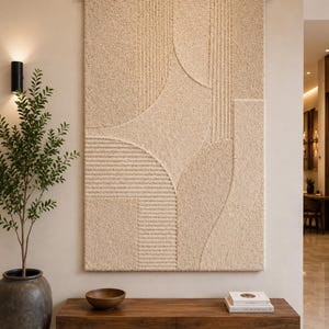 Op de afbeelding: Een grote rechthoekige beige wandhanger van getufte stof met een getextureerd oppervlak, voorzien van uitgesneden geometrische patronen van verticale lijnen en gebogen vormen, opgehangen aan een horizontale houten roede.