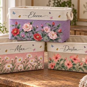 Könnte beinhalten: Drei rechteckige Aufbewahrungskörbe aus Stoff mit Seilgriffen. Der obere Korb ist lila mit roten und rosa Rosen und dem Namen Eleven. Der untere linke Korb ist rosa mit weißen Gänseblümchen und dem Namen Max. Der untere rechte Korb ist beige mit rosa Hibiskus und dem Namen Dustin.