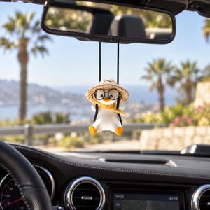 Peut inclure: Une petite figurine de pingouin blanc suspendue au rétroviseur par des cordons noirs. Le pingouin porte un chapeau à texture de paille, des lunettes rondes à monture noire et une écharpe jaune. La figurine a des pieds orange et est assise.