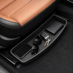 Puede incluir: Una bandeja de almacenamiento de plástico negro para asiento de coche situada en el suelo, que contiene un smartphone negro, gafas de sol negras, una llave de coche plateada con dos llaves metálicas y una cartera rectangular negra junto a un asiento de cuero.