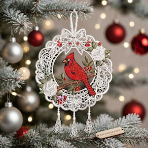 May include: A circular white lace ornament featuring an embroidered red cardinal perched on a brown branch. The lace frame is decorated with white roses, red berries, and a bow at the top. Three white tassels hang from the bottom of the ornament.