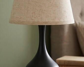Op de afbeelding: Een tafellamp met een matzwarte, druppelvormige voet met een slanke hals en een brede, taps toelopende lampenkap van lichtbeige, gestructureerde stof. De lamp staat rechtop en toont een gladde, effen gekleurde voet en een neutrale kap.