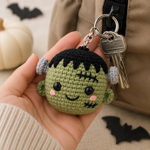 May include: A round, light green crocheted keychain shaped like a monster head with black hair, black safety eyes, a stitched smile, pink cheeks, and two small grey bolts on the sides. The keychain is attached to a silver metal ring with a key.