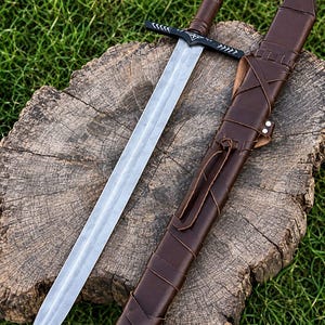 May include: A steel sword with a double-edged blade and a brown leather-wrapped handle. The black crossguard and pommel feature white etched geometric patterns. A matching brown leather scabbard with leather lacing lies beside the sword.