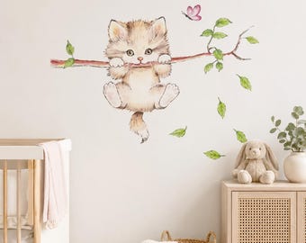 May include: A wall decal featuring a fluffy light brown kitten hanging from a thin brown tree branch with green leaves and a small pink butterfly. The kitten has large dark eyes and white paws. The design is applied to a plain off-white wall surface.