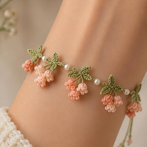 May include: A delicate bracelet featuring crocheted peach-colored flower clusters and green leaves, interspersed with small white pearls. The floral design is crafted from fine thread, creating a botanical pattern that wraps around the wrist.