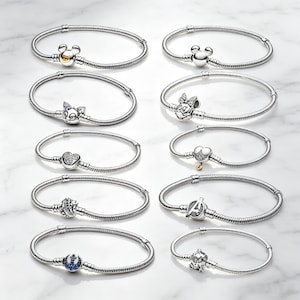 Peut inclure: Dix bracelets en chaîne serpent argentée disposés en deux colonnes. Chaque bracelet présente un charm unique, incluant des motifs inspirés de personnages comme Mickey Mouse, Minnie Mouse, un chat, un cœur, le logo Avengers et un emblème bleu.