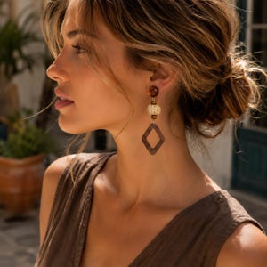 May include: A drop earring featuring a dark brown spherical stud, a small wooden bead, a textured beige woven cylindrical bead, and a dark brown diamond-shaped pendant. The components are connected vertically in a linear arrangement with gold-tone metal hardware.