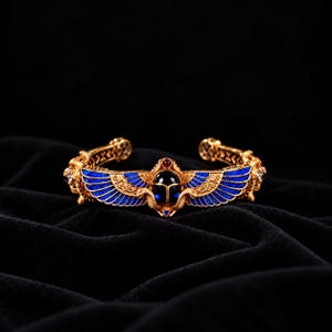 Peut inclure: Un bracelet manchette doré orné d'un scarabée central au corps en pierre sombre, flanqué d'ailes émaillées bleues. Le métal présente des motifs complexes et une petite pierre rouge est sertie au-dessus du scarabée.