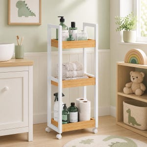 May include: A three-tier rolling storage cart featuring a white metal frame and light wood shelves. The cart holds various bathroom items including bottles, folded towels, and toilet paper rolls. It is equipped with four small white caster wheels.