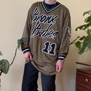May include: An olive green long-sleeve jersey with thin black pinstripes, a V-neck collar, and the text "Bronx Babes" in white script with black outlines. The number "11" is printed in white with black borders on the front, and black pants are worn.