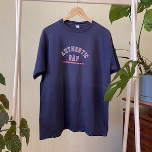 Pode incluir: Uma t-shirt azul-marinho de manga curta com o texto AUTHENTIC GAP WORLDWIDE impresso a vermelho e branco no peito. A peça tem um decote redondo e está pendurada num cabide de madeira, exibindo um design simples e clássico.