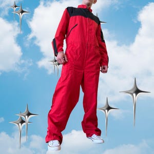 Pode incluir: Um macacão vermelho com um painel preto no peito e nos ombros, fecho de correr frontal completo, mangas compridas com detalhes pretos e gola alta. A peça é usada com ténis brancos sobre um fundo de céu azul com gráficos de estrelas.
