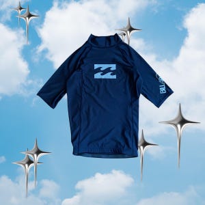 May include: A dark navy blue short-sleeve rash guard shirt featuring a mock neck collar. The chest displays a light blue wave logo, and the left sleeve features the text BILLABONG printed in light blue lettering against the dark fabric.
