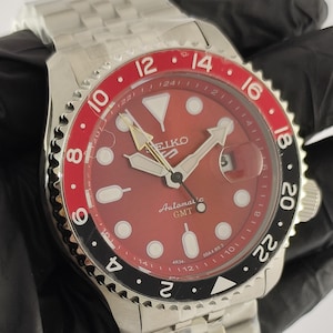 May include: A stainless steel Seiko 5 Automatic GMT wristwatch with a red dial, luminous hour markers, and a date window. The watch features a bidirectional rotating bezel with a red and black insert marked with 24-hour numerals and a metal link bracelet.