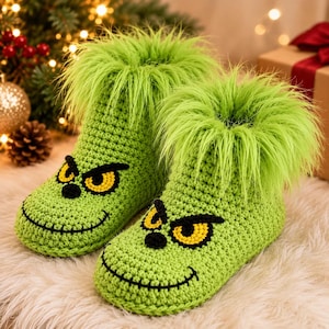 Puede incluir: Un par de pantuflas de ganchillo color verde lima con un borde de piel sintética esponjosa en la parte superior. Las pantuflas tienen ojos amarillos bordados con pupilas negras y una boca negra cosida, formando una cara en la parte frontal.