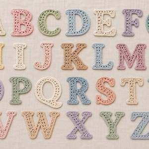 Peut inclure: Un ensemble complet de vingt-six lettres de l'alphabet de A à Z, chacune fabriquée en fil crocheté dans diverses couleurs pastel, notamment crème, rose, bleu clair, vert sauge, lavande et beige, disposées en quatre rangées sur un tissu neutre.