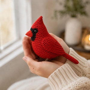 May include: A crocheted cardinal bird figurine featuring a vibrant red body, a pointed crest, a black mask around the eyes, and a small orange beak. The bird is crafted with a textured yarn stitch pattern and has a long, tapered tail.