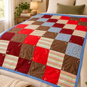 Puede incluir: Una colcha de retazos con una cuadrícula de paneles de tela cuadrados en rojo sólido, granate oscuro, marrón y bloques estampados con flores azules y rayas en zigzag multicolores. La colcha tiene un borde azul y está atada con hilo.