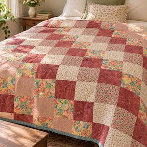 Puede incluir: Una colcha de patchwork con una cuadrícula repetitiva de parches de tela cuadrados. El diseño alterna entre cuadrados de color rojo oscuro sólido y varios cuadrados con estampados florales en tonos rosa, amarillo, verde y azul, con un borde azul fino.