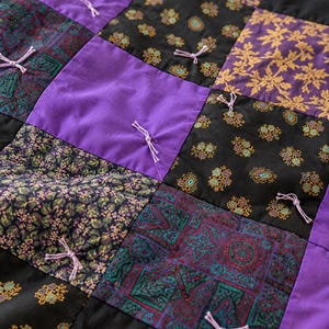 Pu&ograve; includere: Una trapunta patchwork composta da una griglia di pannelli quadrati in tessuto viola tinta unita, nero con motivi a foglie dorate, nero con piccoli motivi floreali e verde ottanio scuro con disegni geometrici, fissati con nodi di filo viola.
