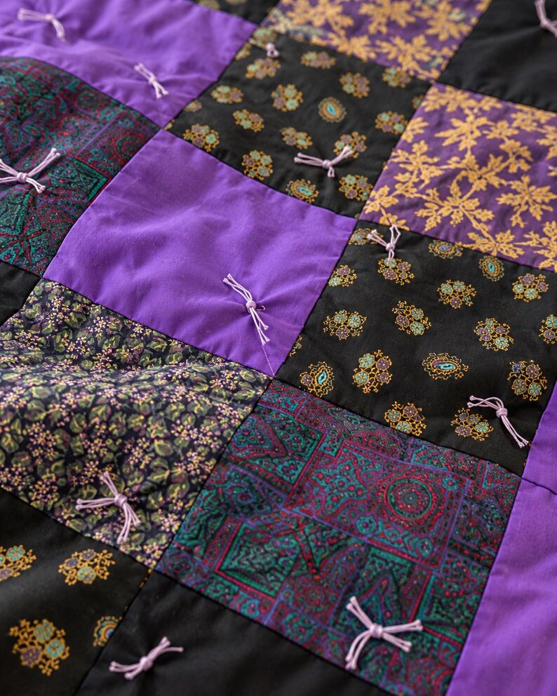 Pu&ograve; includere: Una trapunta patchwork composta da una griglia di pannelli quadrati in tessuto viola tinta unita, nero con motivi a foglie dorate, nero con piccoli motivi floreali e verde ottanio scuro con disegni geometrici, fissati con nodi di filo viola.