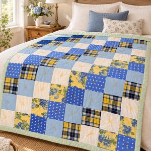 Pode incluir: Uma colcha de retalhos com uma grade de blocos de tecido quadrados em padrões alternados de bolinhas azuis e brancas, estampas florais amarelas e azuis, e xadrez azul-marinho, amarelo e branco, com uma borda verde-clara e laços amarelos.