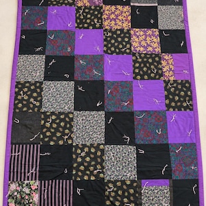 Pu&ograve; includere: Una trapunta patchwork rettangolare caratterizzata da una griglia di quadrati viola a tinta unita alternati a vari quadrati di tessuto fantasia in nero, viola e giallo. La trapunta &egrave; rifinita con un bordo viola e annodata con fili bianchi.