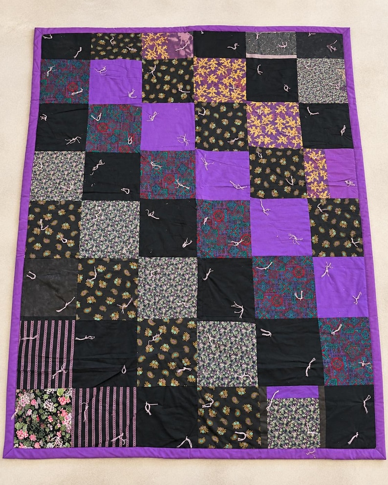 Pu&ograve; includere: Una trapunta patchwork rettangolare caratterizzata da una griglia di quadrati viola a tinta unita alternati a vari quadrati di tessuto fantasia in nero, viola e giallo. La trapunta &egrave; rifinita con un bordo viola e annodata con fili bianchi.