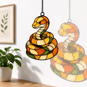 May include: A stained glass suncatcher shaped like a coiled snake, featuring segments of orange, yellow, cream, and green glass held together by black metal cames. The snake is suspended from a black metal chain for hanging display.