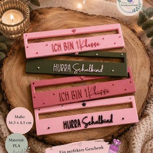 Könnte beinhalten: Vier rechteckige Schilder mit den Maßen 16,5 x 4,5 cm, jeweils mit einem horizontalen Schlitz und erhabenem Text. Zwei Schilder sind rosa mit dem Text "ICH BIN 1. Klasse", eines ist olivgrün mit "HURRA Schulkind" und eines ist dunkelrosa mit "ICH BIN 1. Klasse".