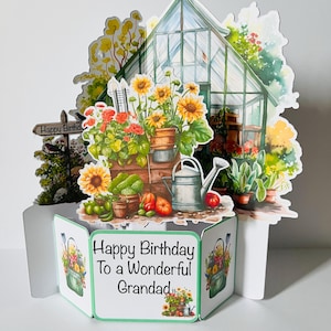 May include: A pop-up birthday card depicting a garden scene with a greenhouse, sunflowers, and a watering can. The card reads "Happy Birthday to a Wonderful Grandad" with floral illustrations. The card is white with green borders.