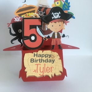 May include: A red and yellow pop-up card with a pirate theme. The card features a pirate holding a sword, a treasure chest, an octopus, a parrot, a ship, and the number 5. The card also has the text "Happy Birthday Tyler" on it.