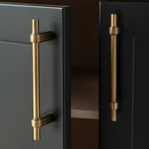 Op de afbeelding: Twee verticale messing kastgrepen met een cilindrische stang en getrapte montagebeugels bevestigd aan matzwarte kastdeuren. Het logo van Golden Home Hardware is zichtbaar in de linkerbovenhoek van de afbeelding met een rond icoon.