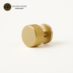 May include: A cylindrical brass cabinet knob with a brushed finish and a textured, cross-hatched knurled edge. The knob features a smooth, flat circular front face and a circular base plate. The text GOLDEN HOME HARDWARE is visible.