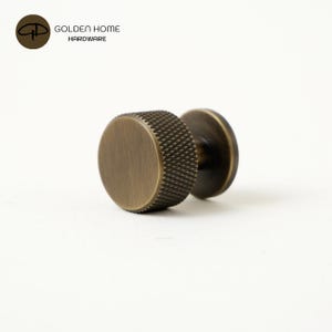 May include: A cylindrical cabinet knob with a brushed antique brass finish. The knob features a flat circular face and a textured, knurled edge pattern around the circumference. It is attached to a circular base plate, shown from a side angle.