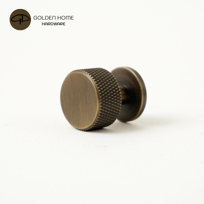 May include: A cylindrical cabinet knob with a brushed antique brass finish. The knob features a flat circular face and a textured, knurled edge pattern around the circumference. It is attached to a circular base plate, shown from a side angle.