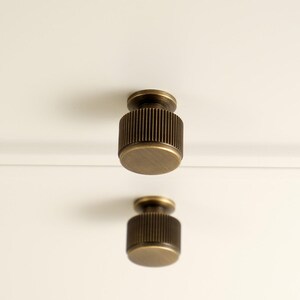 May include: Two cylindrical cabinet knobs with a brushed antique brass finish and vertical ribbed detailing along the sides. The knobs are mounted on a plain white surface. The logo GOLDEN HOME HARDWARE is visible in the top left corner.