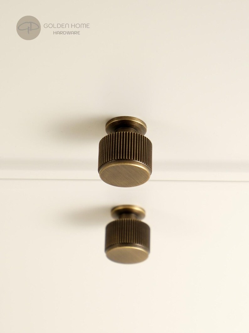 May include: Two cylindrical cabinet knobs with a brushed antique brass finish and vertical ribbed detailing along the sides. The knobs are mounted on a plain white surface. The logo GOLDEN HOME HARDWARE is visible in the top left corner.