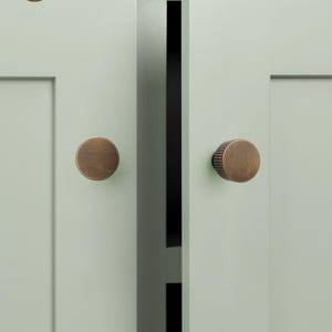 Op de afbeelding: Twee ronde kastknoppen van geborsteld brons met verticaal geribbelde details aan de zijkanten, gemonteerd op licht saliegroene kastdeuren in shaker-stijl. Het logo GOLDEN HOME HARDWARE is zichtbaar in de linkerbovenhoek van de afbeelding.