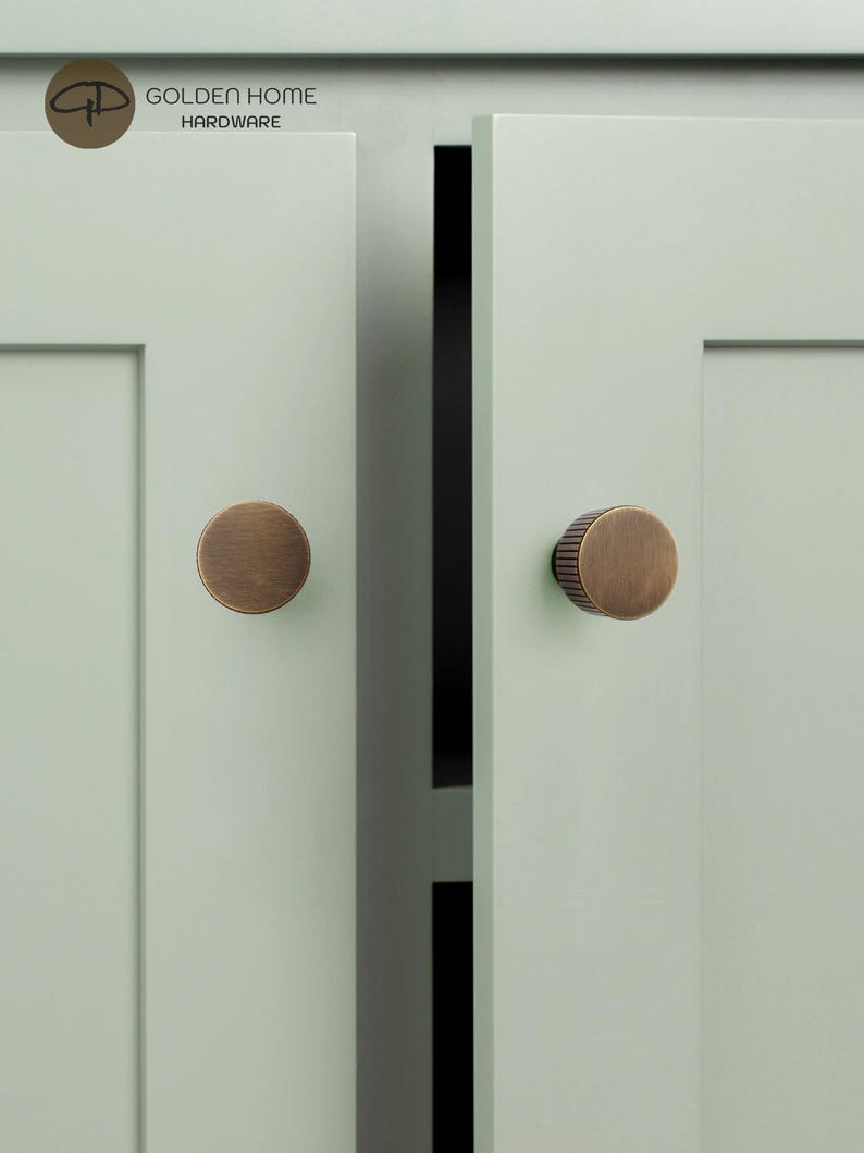Op de afbeelding: Twee ronde kastknoppen van geborsteld brons met verticaal geribbelde details aan de zijkanten, gemonteerd op licht saliegroene kastdeuren in shaker-stijl. Het logo GOLDEN HOME HARDWARE is zichtbaar in de linkerbovenhoek van de afbeelding.
