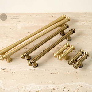 May include: A collection of brushed brass cabinet hardware including two long bar pulls, one medium bar pull, and two small T-bar knobs. Each piece features a cylindrical rod design with decorative finial ends and a logo reading GOLDEN HOME HARDWARE.