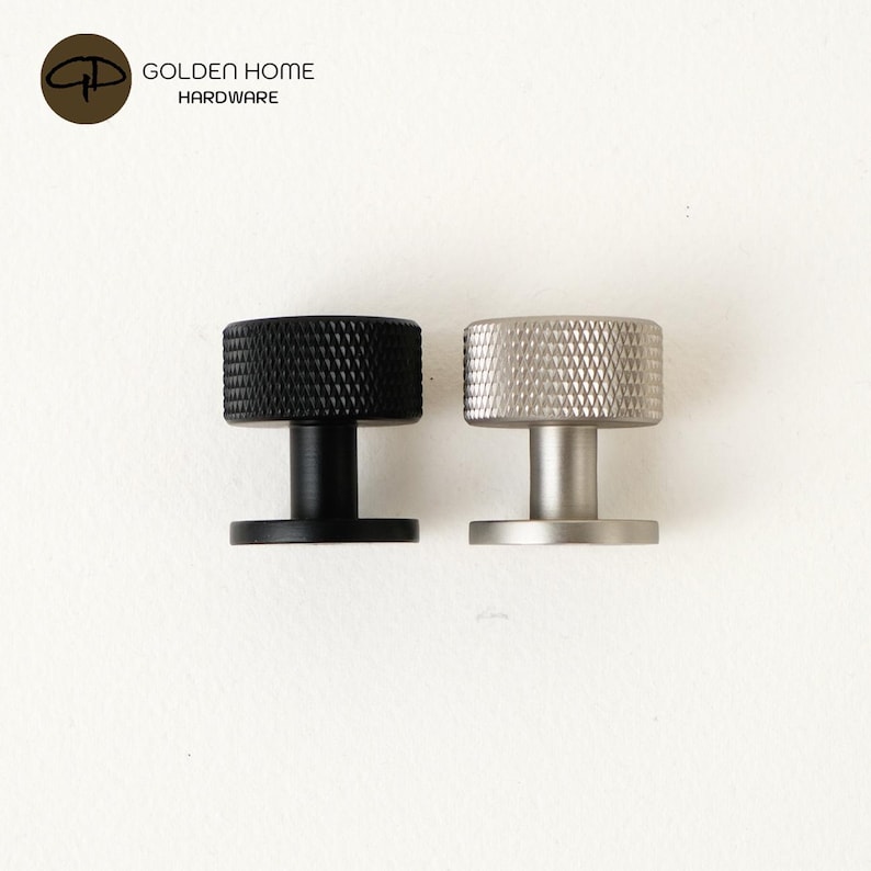 May include: Two cylindrical cabinet knobs, one matte black and one brushed silver, featuring a textured diamond-patterned knurled edge on the top surface and a smooth base. The logo GOLDEN HOME HARDWARE is visible in the top left corner.