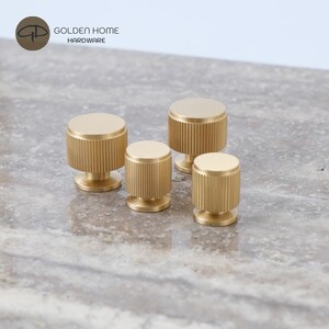 May include: Four cylindrical cabinet knobs made of brushed gold metal, each featuring a flat top and a vertically ribbed, textured side surface. The knobs are arranged in a cluster, showcasing a consistent metallic finish and uniform design.