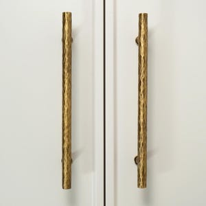 May include: Two vertical brass cabinet pulls with a hammered texture mounted on white double doors. The hardware features a cylindrical shape and a metallic gold finish. The logo for Golden Home Hardware is visible in the top left corner.